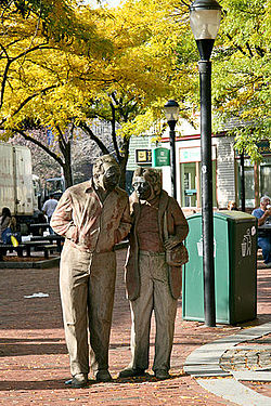 A sculpture resembling an elderly man and woman walking down the street with trees and buildings in the background.