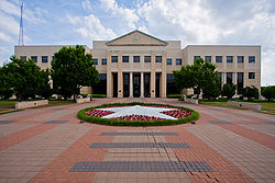 Front picture of a two-story administration building on a cloudy day. The walkway is shown leading up to the building including a circular garden in with white flowers forming a star.