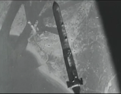 View from a high-altitude aircraft, looking down and showing a bomb that has just being released. On the ground can be seen a box-like building next to water