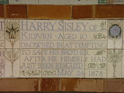 A tablet formed of five tiles of varying sizes, bordered by yellow and blue flowers in an art nouveau style. The tablet reads "Harry Sisley of Kilburn aged 10 drowned in attempting to save his brother after he himself had just been rescued May 24, 1878".