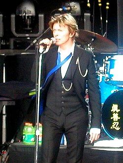 A male singer wearing a dark grey three-piece suit, white shirt and a blue undone tie, on stage singing while holding a microphone on a stand.