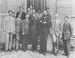 Fifteen men in suits, and one womyn, pose for a group photograph