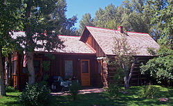 An unpainted dark brown wooden house amid tall trees in two sections. The taller one, on the right, has a pointed roof with a small brick chimney rising from the middle.