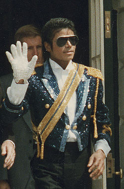 A mid-twenties Michael Jackson wearing a sequined military jacket and dark sunglasses. He is walking while waving his right hand, which is adorned with a white glove. His left hand is bare.