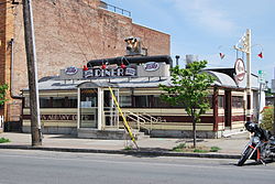 A single-storey building in the shape of a railroad car, with an Art Deco facade in cream and maroon stripes situated next to a taller brick building