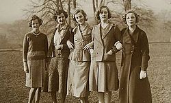 Nearly full length group portrait of five well-dressed women standing in a field. Their ages range from roughly 20 to 30; their hair is cut short of the shoulders in elegant 1930s or 1940s styles; four of the five wear skirts down just below the knee, and one a longer coat. Two wear pearls.