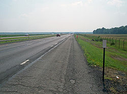 An expressway, seen from the shoulder of its righthand lanes, continues straight ahead to the center of the image. Either side is fenced off; the surrounding area is mostly marsh with a wooded area visible at right center