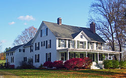 A large white house with a black pointed roof, front porch and rear wings seen from downhill and slightly to its left. It is shaded by a tall tree on the right.