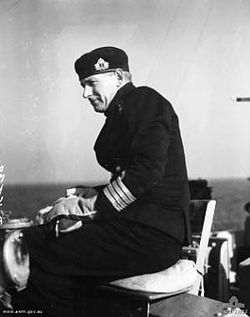 A man in naval uniform sitting on a seat on the bridge of a ship. He is wearing think gloves and a cap, and is slightly side on to the camera. A body of water can be seen in the background.