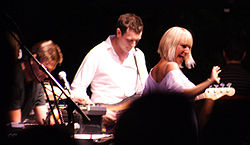 Upper body shot of three musicians performing. Left man is seen in right profile facing downwards and partly obscured by equipment. Second man is playing a guitar and is looking down. Furler is shown from behind, she is partly turned to her right, smiling and gesturing with her right arm. She is partly obscured by an audience member's silhouette.