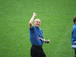 A middle-aged man. He is wearing a blue polo shirt and black trousers, and standing on a grass field.