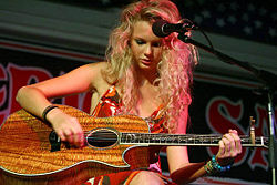 A young female with curly blond hair faces down at an acoustic guitar made of koa wood while a large microphone is placed close to her. She is wearing patterned, red dress.