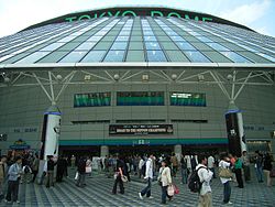 A large circular, white building; a slanted, glass overhang protects people walking around the plaza in front of the building's entrance below. A large banner above the entrance reads "ROAD TO THE NIPPON CHAMPIONS".