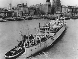 The USS Chaumont, a large light-coloured transport vessel with a smaller vessel alongside, against a background of shorefront buildings and docked ships