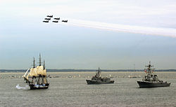 Photo of a ship at sail with two escorts as navy jets fly overhead