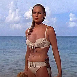 A blonde woman wearing a white bikini and a belt, on which a knife can be seen.