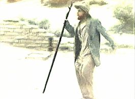 Still film image showing a bearded man wearing a jacket, shirt, and pants bulging at the thighs, with apparently normal leather shoes, carrying a walking staff