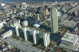 An aerial view of a granite plaza with multiple pools surrounded by four identical marble, steel, and glass buildings toward the bottom and one large steel and glass building near the top of the view. Surrounding these main buildings is a cityscape.