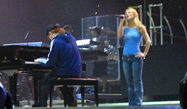 Image of a blond female performer. She is wearing blue jeans and a sleeveless light blue top. She is standing next to a piano player, who is playing a big black piano. Her left hand is in her hip while her right hand holds her mic while she is singing. Two members of the crew stand a few meters behind them.