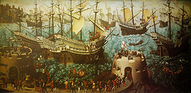 A small fleet of large, highly decorated carracks are riding on a wavy sea. In the foreground are two low, fortified towers bristling with cannons and armed soldiers and retinue walking between them.