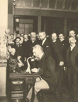 A grizzly and well-dressed Alexander Graham Bell sits at a desk talking over a candle-stick telephone, surrounded by numerous business executives and news reporters who are witnessing a historic event in the atrium of a large corporate building.