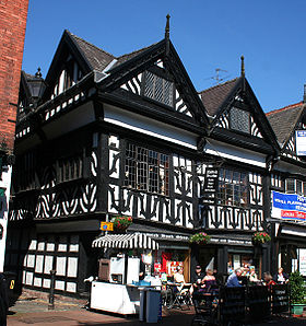 A black-and-white building in a corner position, with two gables to the front and one visible to the left side. On the street in front is a pavement café.