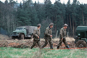 Three uniformed men, armed with assault rifles, walking in a column through a grassy landscape with dense trees in the background. A canvas-sided truck is visible in the left background and part of another vehicle is seen on the right in a stretch of ploughed-up ground.