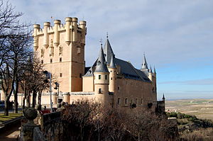 A castle high on a rocky peninsula above a plain. It is dominated by a tall rectangular tower rising above a main building with steep slate roof. The walls are pink, and covered with a sculptural pattern. There is a variety of turrets and details.