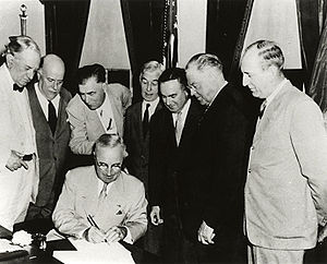 A man in a suit is seated at a desk, signing a document. Seven men in suits gather around him.