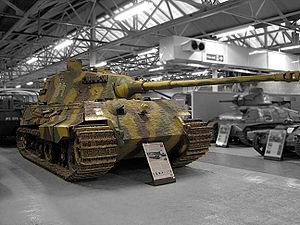 A large, turreted tank with dull yellow, green and brown wavy camouflage, on display inside a museum. The tracks are wide, and the frontal armor is sloped. The long gun overhangs the bow by several meters.