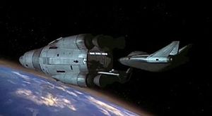 A pair of spacecraft, both designed to a curved aesthetic, orbit an Earth-like planet. One vessel, smaller than the other, is a shuttle departing from the docking port of the mothership.