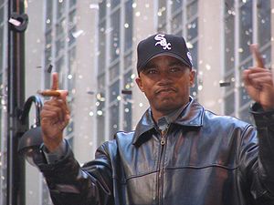 A dark-skinned man in a black baseball cap and black leather jacket holding up both his hands with his index fingers extended. He is holding a cigar in his right hand.