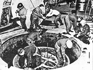 Soldiers and workmen, some wearing steel helmet, clamber over what looks like a giant manhole.