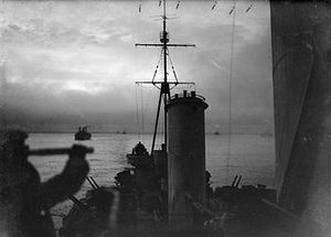 View from the cruiser HMS Sheffield as she sails on convoy duty through the waters of the Arctic Ocean. In the background are merchant ships of the convoy. The image was taken during the twilight of the arctic winter—the short time each day that the sun is seen during winter near the pole. In the foreground is the silhouette of a lookout using a telescope.