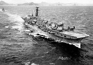 A small aircraft carrier travelling at speed through choppy water. Several propeller aircraft with folded wings are sitting on the flight deck. A small warship is following in the carrier's wake, and a rocky, hilly shoreline can be seen across the horizon