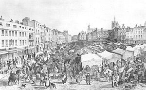 Very crowded market on a slope. At the left of the picture is a row of tall thin shops, and behind the market is another row of shops surrounding a tall church.