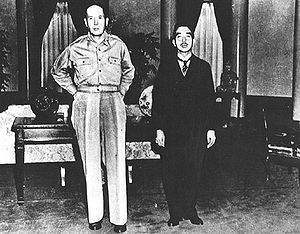 MacArthur, without hat and wearing open necked shirt and trousers, towers over an oriental man in a dark suit.