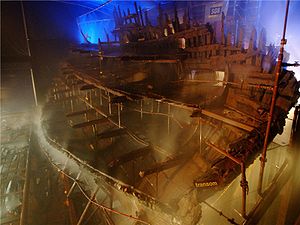 A diagonal section of a wooden ship seen from the stern inside a moodily lit building while it is being sprayed with water from a sprinkler system