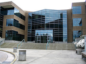 A brown building stretching from the extreme left to the extreme right and displaying many large windows reflecting daylight. The sky above the building is white and the ground in the foreground is grey concrete.
