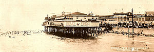 A black-and-white postcard shows a photograph, taken from a location on the water, of a large building sitting on pier by the beach. The beach is fronted by a seawall and a crowded waterfront beyond. The caption on the postcard says "Where life is worth living at Galveston".