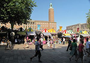 Row of brightly coloured market stalls. Behind the market stalls is a very large red brick building with a tall clock tower. Next to it is a long low dark stone building.