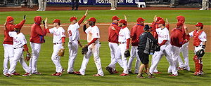 A group of men in white baseball uniforms with red pinstripes and red baseball caps high-five each other while passing in lines moving in opposite directions.
