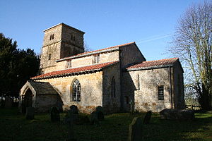 A stone church with a red tiled roof seen from the southeast, with a small chancel, a larger nave with clerestory and porch, and a tower