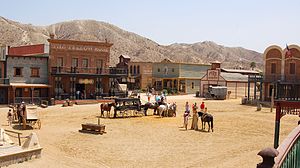 Squarish buildings made of wood and bricks surround a square, where horses and carriages gather.