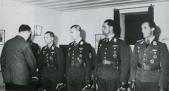 Five men all wearing military uniforms and decorations standing in row. The man on the far left is shaking hands with another man whose back is facing the camera.