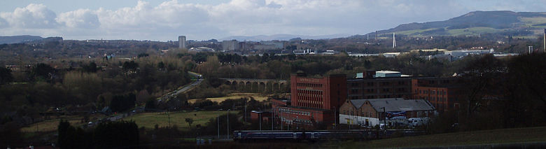 View of Glenrothes seen in its landscape setting from a nearby cemetery. A train is leaving nearby Markinch Station on the East Coast Mainline. Glenrothes town centre with the numerous taller residential and office buildings can be seen in the centre of the image. The River Leven Bridge provides a stark white vertical emphasis on the right side of the image. The Lomond Hills regional park and rolling countryside form the backdrop on the horizon