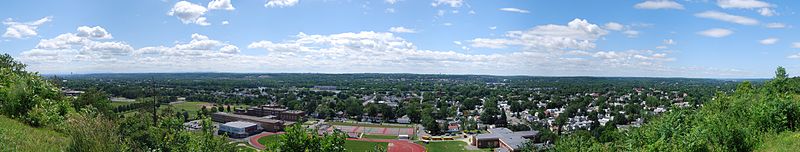A wide panoramic shot begins and ends in bushes but is graced by two and three story city houses throughout most of the image. Front and center are three large schools featuring a track, tennis courts, and a football field. Behind the houses is a river flowing right to left.