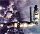 A collection of three space station modules, arranged in an L-shape. At the left of the image is a Soyuz spacecraft docked to a short, stubby module, in turn docked to a larger, stepped-cylindrical module covered in white insulation, from which solar arrays project. A third module, similar in size to the stepped-cylinder module, is attached to the upper port of that module, forming the up-stroke of the L. This module is also projecting two solar arrays, and the cloudy Earth serves as the image's backdrop.