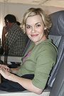 A woman with short hair in an airplane seat wearing a green sweatshirt and smiling